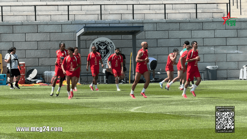 الحصة التدريبية للمنتخب الوطني بعد مباراة البرازيل Soccer players in red uniforms running on a training field near a stone wall with a team logo backdrop.