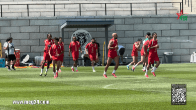 Soccer players in red uniforms running on a training field near a stone wall with a team logo backdrop.