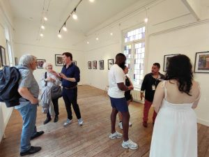 People mingle in a bright white-walled art gallery with framed pictures on the walls and wooden floors during an event.