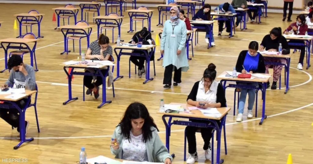 WhatsApp Image 2026-06-05 at 135531 | MCG24 | موقع إلكتروني مغربي شامل متجدد على مدار الساعة Exam session in a gym: students at single desks spaced apart, supervisor in a light blue coat wearing a mask.