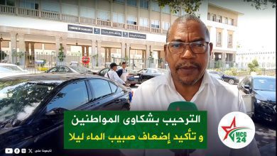 Male reporter wearing glasses speaks to camera outside a government building, with a green Arabic banner at the bottom and parked cars behind him.