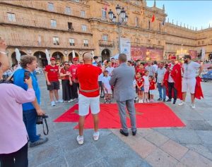 Group of people in red shirts standing on a red carpet in a historic plaza while a man in a gray suit speaks to them.