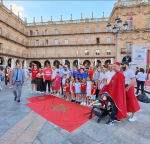 Group of children and adults posing in a historic plaza with a red Moroccan flag spread on the ground in front of them.