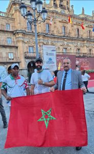 Three men hold a red Moroccan flag with a green star in a historic plaza, with flags and banners on an ornate building behind them.