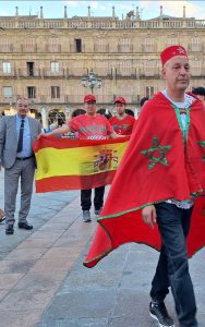 Group of people in a city square holding a large Spanish flag, with others in red shirts nearby and one man in Moroccan-inspired red cape and fez-style hat as part of a procession.