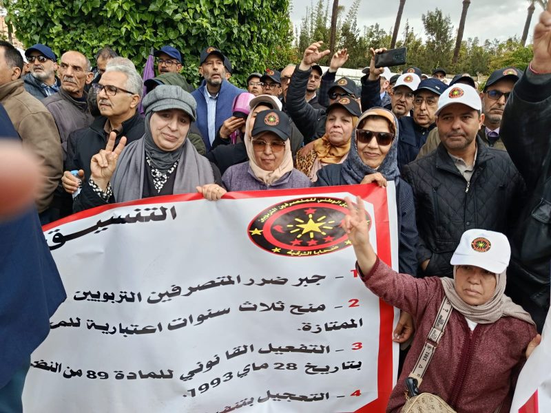 Crowd of protesters holding a banner with Arabic text and a circular emblem; several people raise their hands, some making peace signs.
