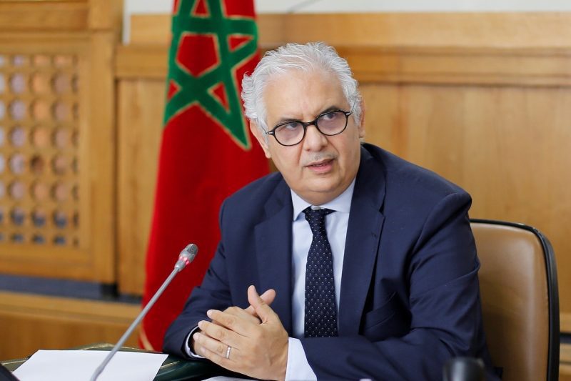 WhatsApp Image 2026-06-19 at 132921 | MCG24 | موقع إلكتروني مغربي شامل متجدد على مدار الساعة Middle-aged man with gray hair and glasses in a navy suit speaks into a microphone at a formal meeting, Moroccan flag in the background.