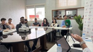 WhatsApp Image 2026-06-21 at 185258 | MCG24 | موقع إلكتروني مغربي شامل متجدد على مدار الساعة A group of diverse people seated around a long conference table listening to a speaker, with a projector on the table and a green microphone labeled MCG24 nearby.