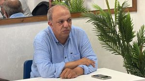 WhatsApp Image 2026-06-21 at 185259 2 | MCG24 | موقع إلكتروني مغربي شامل متجدد على مدار الساعة Man in a light blue button-down shirt sits at a white table, hands folded, with a smartphone on the table and green plants in the background.