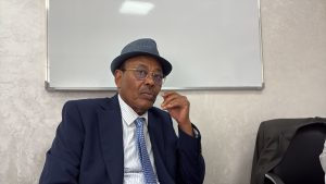 WhatsApp Image 2026-06-21 at 185301 3 | MCG24 | موقع إلكتروني مغربي شامل متجدد على مدار الساعة Older man in a navy suit and blue hat sits in a conference room, hand near his chin with a thoughtful expression, whiteboard behind him.