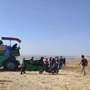 Group of people gathered around a large green combine harvester in a dry field under a clear blue sky.