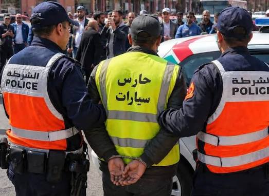 images 43 | MCG24 | موقع إلكتروني مغربي شامل متجدد على مدار الساعة Police detain a man in a crowd; two officers flank him, one in a bright pink-and-orange vest and another in navy, with a yellow vest reading 'حارس سيارات' (parking attendant).
