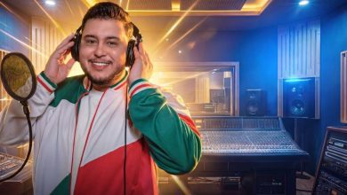 Smiling man wearing a green, white, and red jacket tunes a microphone in a professional recording studio with headphones on.
