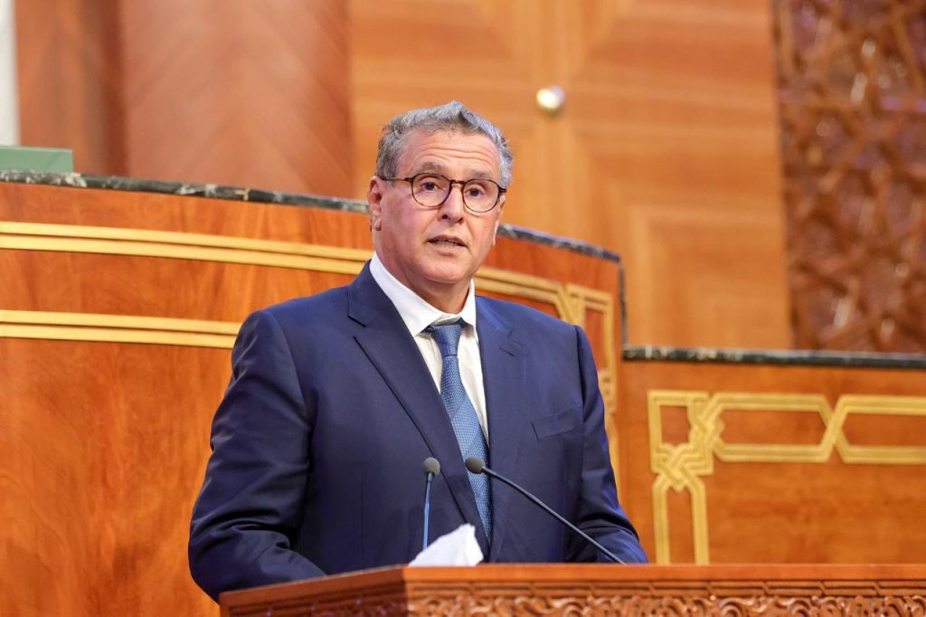 Man in a dark blue suit and glasses delivering a speech at a wooden podium with microphones in a formal setting