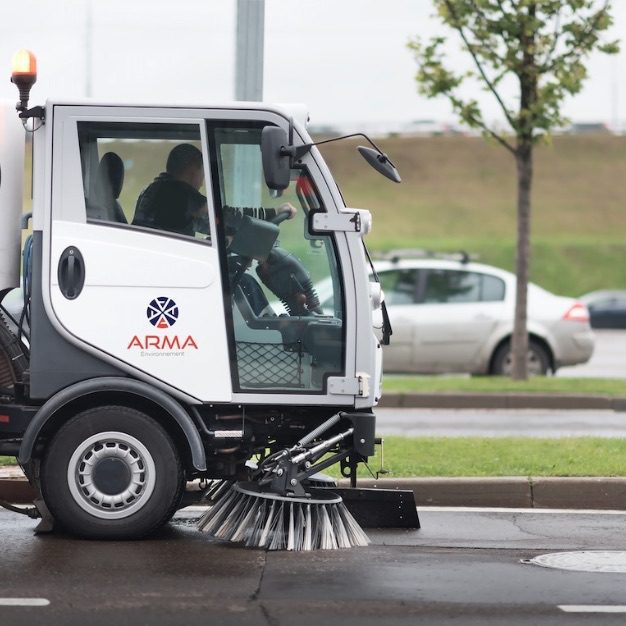 s1 | MCG24 | موقع إلكتروني مغربي شامل متجدد على مدار الساعة White street-sweeping truck in operation, brushing the road surface with a large undercarriage broom, driver visible inside the cab (ARMA Environmental logo on the side).