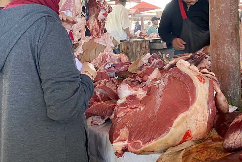 Meat market stall with large cuts of beef on a table; a customer in a gray hoodie selects meat while others shop in the background.