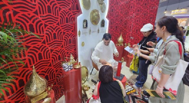 Man in white arranges gold offerings on a small altar as three customers observe, in a shop with bold red patterned walls and ornate decorative items.