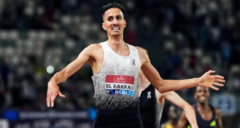 vis_0601202600241904 | MCG24 | موقع إلكتروني مغربي شامل متجدد على مدار الساعة Male sprinter in a white-to-gray sleeveless top with bib El Bakkali Rabat, arms outstretched in celebration on a track.