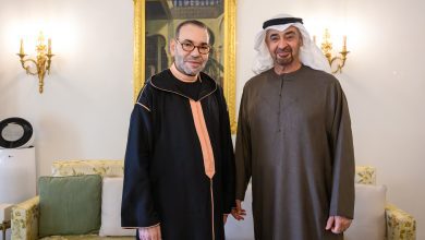 Two men in traditional robes stand side by side in a living room with a gold-framed painting behind them and ornate wall lamps on either side.