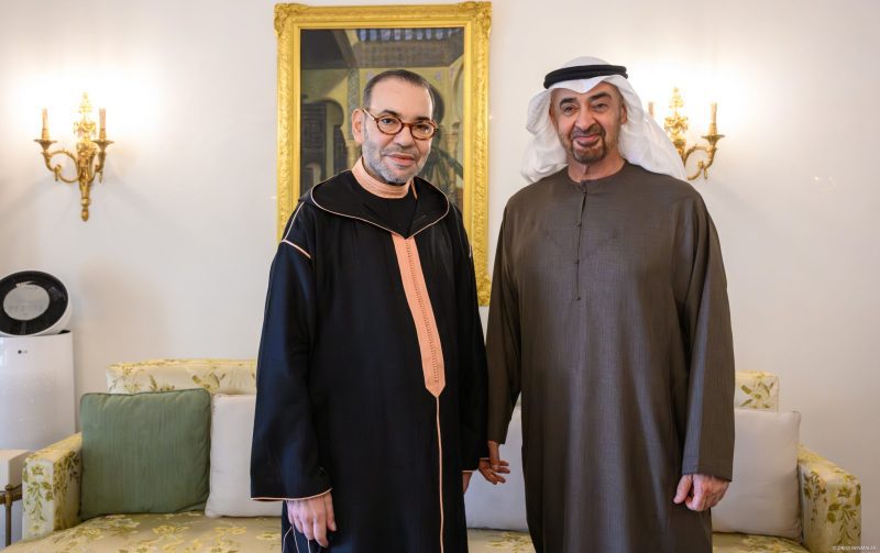 vis_0602202621404400 | MCG24 | موقع إلكتروني مغربي شامل متجدد على مدار الساعة Two men in traditional robes stand side by side in a living room with a gold-framed painting behind them and ornate wall lamps on either side.
