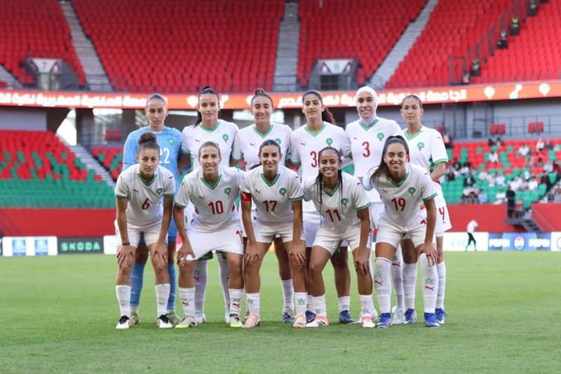 vis_0605202622403136 | MCG24 | موقع إلكتروني مغربي شامل متجدد على مدار الساعة Group of female soccer players in white jerseys with green trim posing for a team photo on a soccer field inside a stadium with red and green seating; goalkeeper in blue stands at left back.