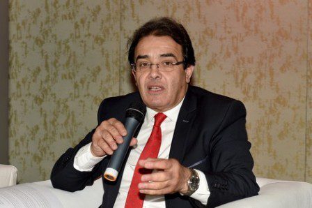 Man in a dark suit and red tie speaks into a handheld microphone while seated on a white couch during a discussion.