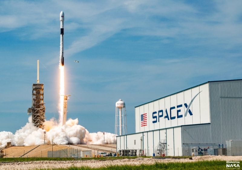 سبيس-إكس | MCG24 | موقع إلكتروني مغربي شامل متجدد على مدار الساعة SpaceX rocket lifting off from a launch pad beside a large SpaceX building, with smoke and a bright flame plume behind it.