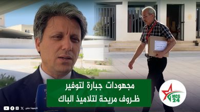 Video thumbnail: reporter in foreground filming outdoors as a man walks with a parcel near a building; green Arabic caption overlay and logos visible.