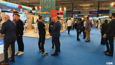 People in business attire talking at a bustling trade show with booths and banners in the background, blue carpet underfoot.