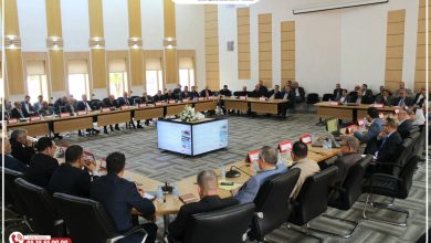 Large conference room with many attendees in suits seated around a U-shaped table, a central screen showing slides, and a decorative banner/logo at the front.