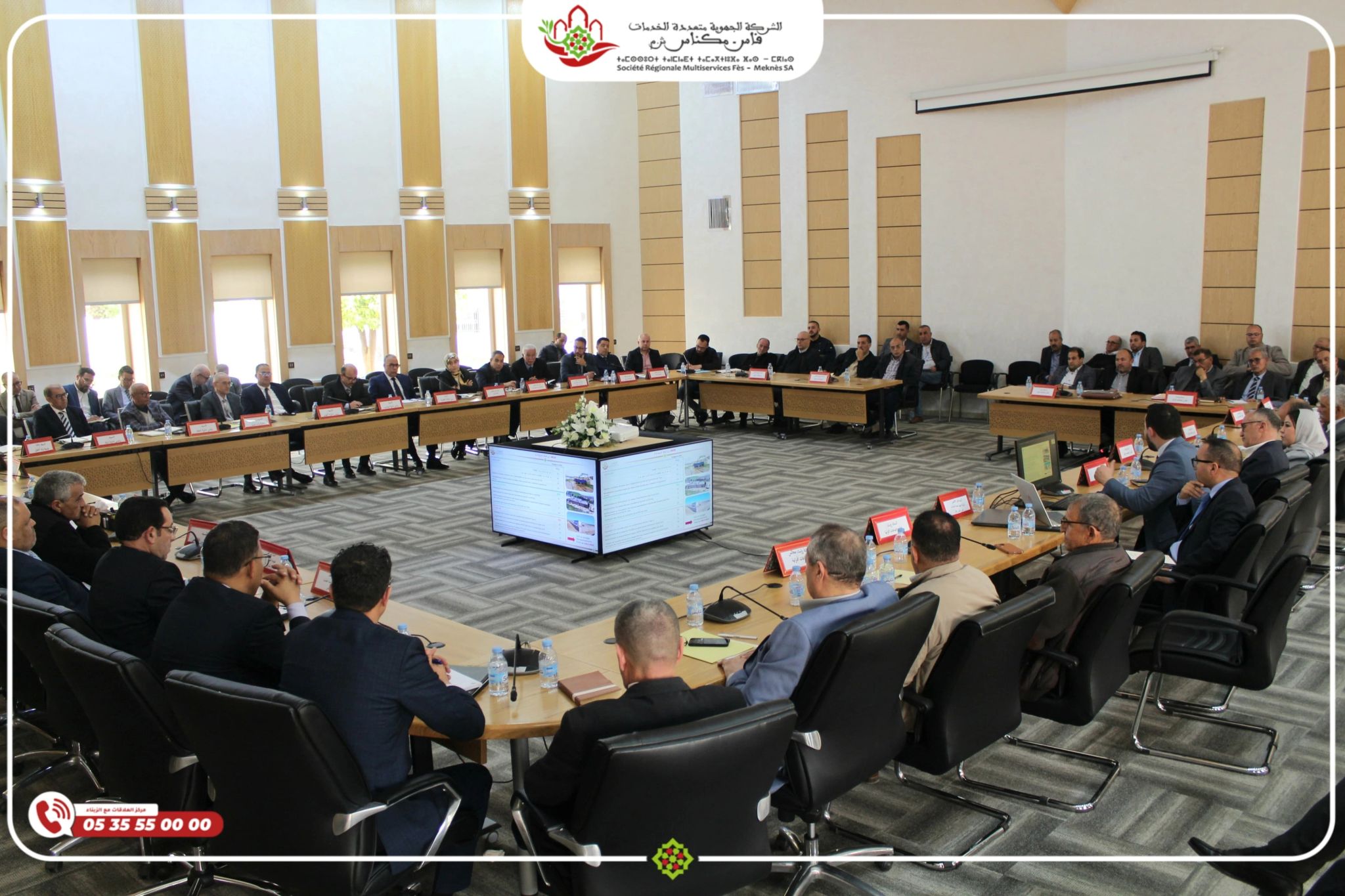 Large conference room with many attendees in suits seated around a U-shaped table, a central screen showing slides, and a decorative banner/logo at the front.