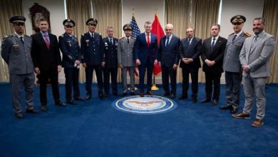 Group of military officers and officials posing for a ceremony photo in front of flags and a blue carpet seal.