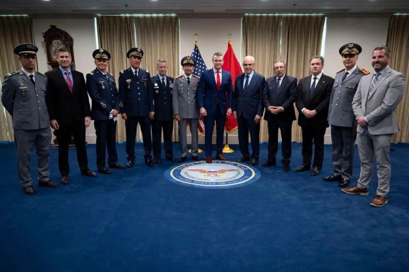 Group of military officers and officials posing for a ceremony photo in front of flags and a blue carpet seal.