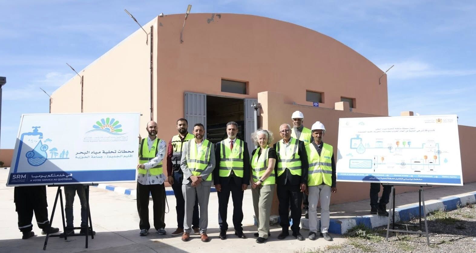 Group of workers and engineers in high‑visibility vests pose in front of a water treatment facility with informational boards beside them.