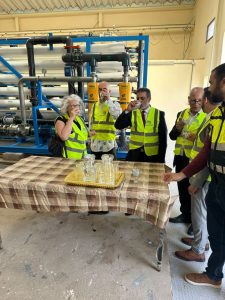 Group of six people in high-visibility vests raising glasses in a toast beside an industrial filtration system.