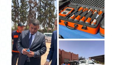 Group of professionals outdoors, a man in a suit examines a small blue device while two colleagues look on in the background.