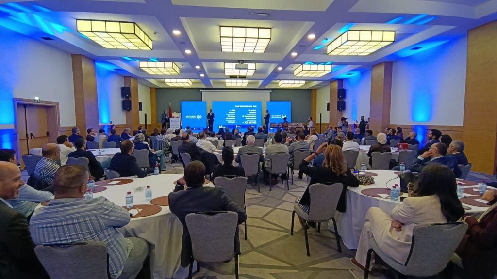 1777201707547 | MCG24 | موقع إلكتروني مغربي شامل متجدد على مدار الساعة Large conference room filled with professionals seated at round tables facing a panel on a stage with three blue display screens.