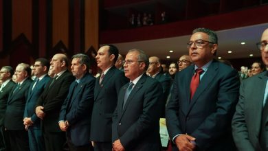 Group of men in dark suits standing in a formal ceremony, hands folded in front, facing forward in a dimly lit auditorium.