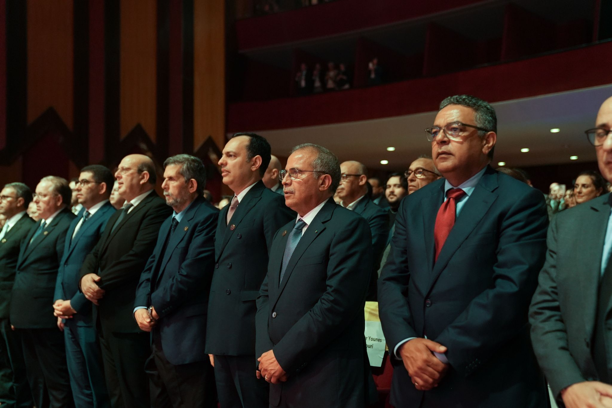 Group of men in dark suits standing in a formal ceremony, hands folded in front, facing forward in a dimly lit auditorium.