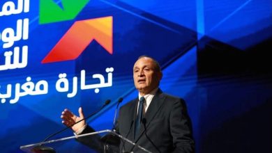 Man in a dark suit giving a formal speech at a transparent podium on a stage with a blue backdrop and Arabic text/logo behind him.