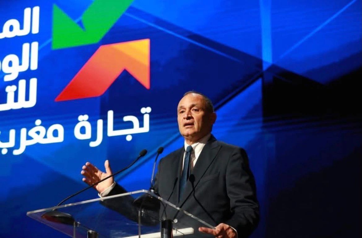 Man in a dark suit giving a formal speech at a transparent podium on a stage with a blue backdrop and Arabic text/logo behind him.