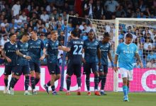 Soccer players in dark blue jerseys celebrate near the goal as teammates congratulate each other, with a light-blue-clad goalkeeper walking away in the foreground.