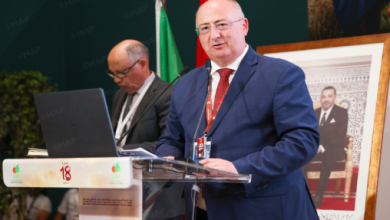 Man in a blue suit and red tie speaks at a clear podium during a formal conference, with a laptop and a secondary speaker behind him.