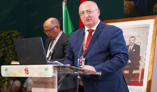Man in a blue suit and red tie speaks at a clear podium during a formal conference, with a laptop and a secondary speaker behind him.