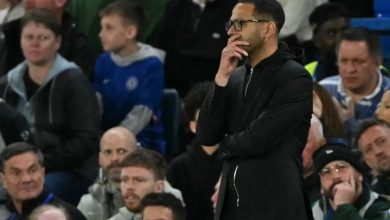 Man in a black coat and glasses stands with his hand on his chin amid a crowded stadium audience, watching an event.