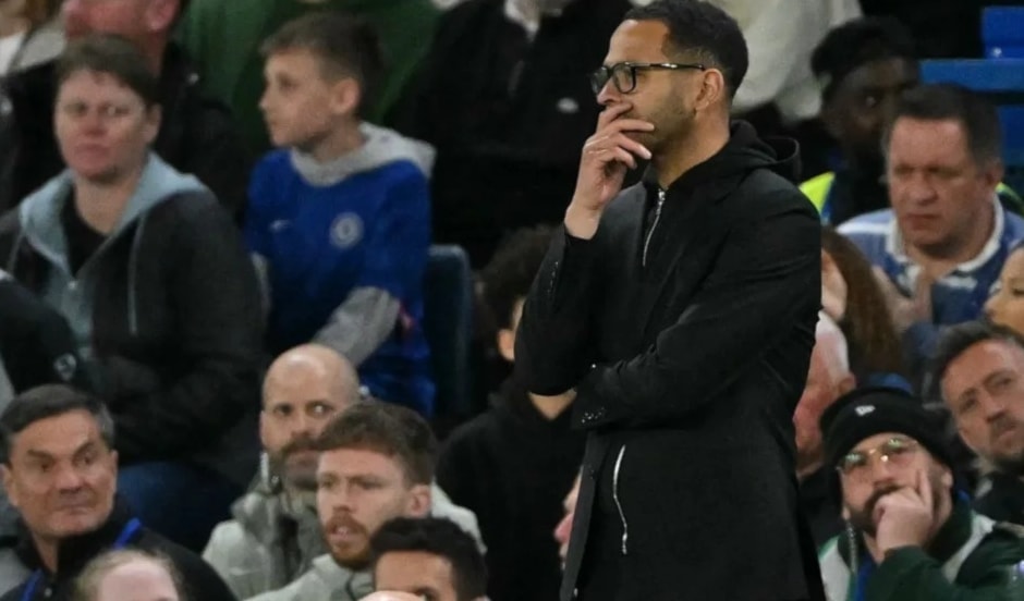 Man in a black coat and glasses stands with his hand on his chin amid a crowded stadium audience, watching an event.
