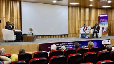 Panel discussion on a stage with three speakers seated in chairs, an empty screen behind them, and an audience in red seats watching.