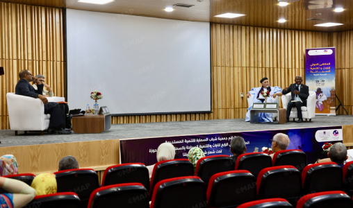 Panel discussion on a stage with three speakers seated in chairs, an empty screen behind them, and an audience in red seats watching.