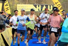 Group of runners sprint toward the finish on a blue carpet, bib numbers visible, with cheering spectators and yellow sponsor banners in the background.