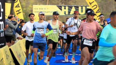 Group of runners sprint toward the finish on a blue carpet, bib numbers visible, with cheering spectators and yellow sponsor banners in the background.